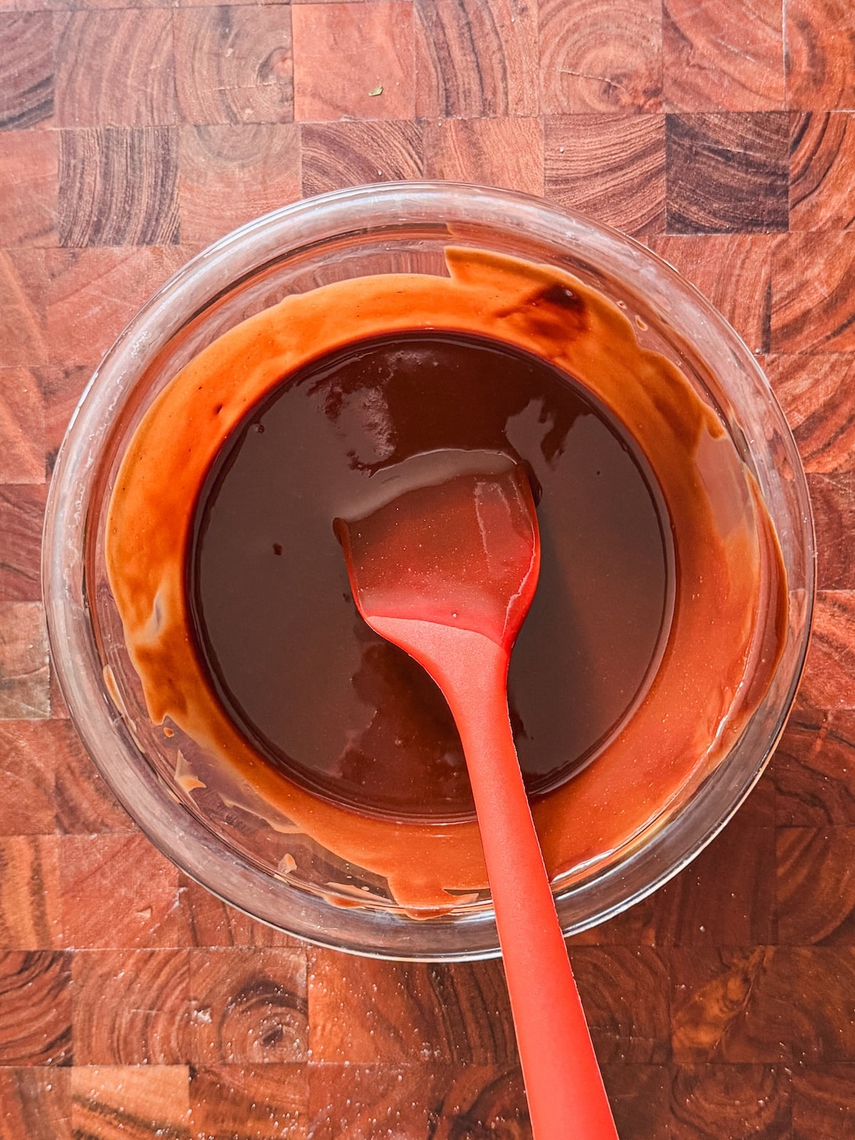 A glass bowl filled with chocolate mixture sits on a wooden surface. A red silicone spatula, coated in the mixture, rests inside the bowl.