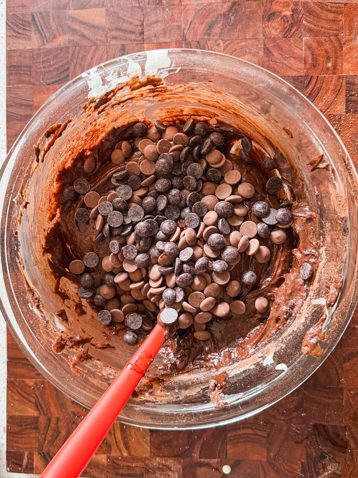 A glass bowl filled with brownie batter and topped with dark and milk chocolate chips sits on a wooden surface. A red spatula is partially submerged in the mixture, ready for stirring.