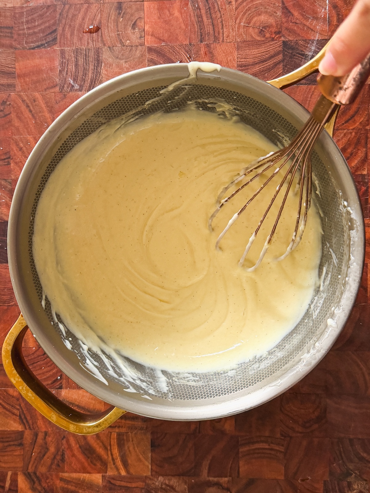 A hand holding a whisk stirs a creamy, light-colored pudding in a metal pan with gold handles, set on a wooden surface.