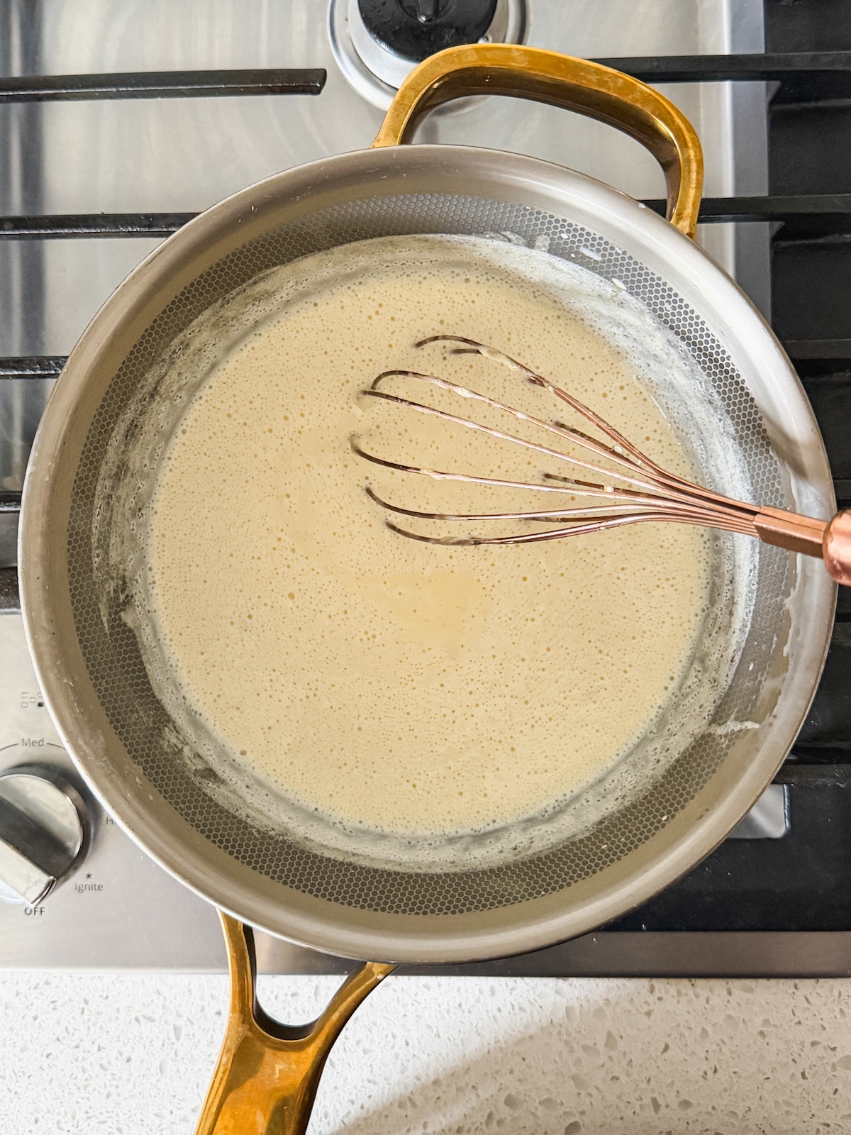 A metal pan with pudding being whisked on a stovetop. The pan has gold handles, and the sauce appears smooth and creamy, with some bubbling at the edges.