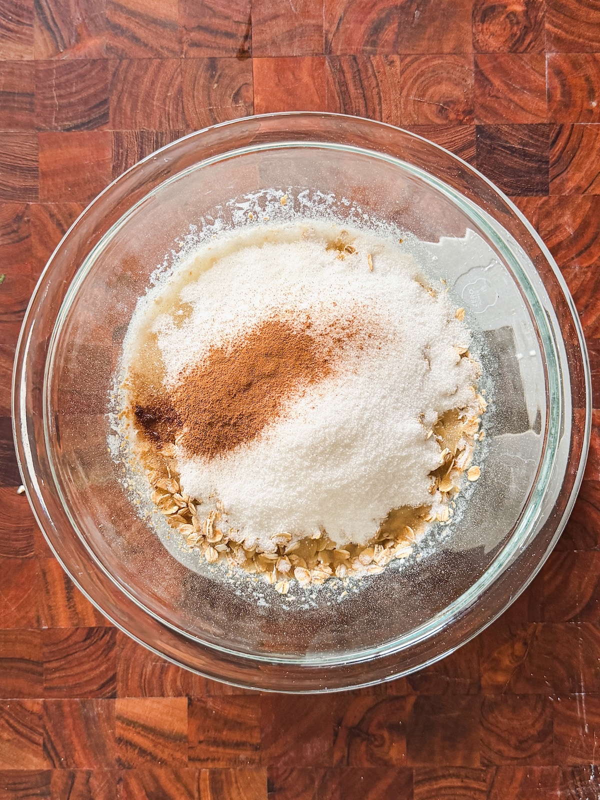 A glass bowl on a wooden surface filled with oats, sugar, cinnamon, and other baking ingredients, ready to be mixed.