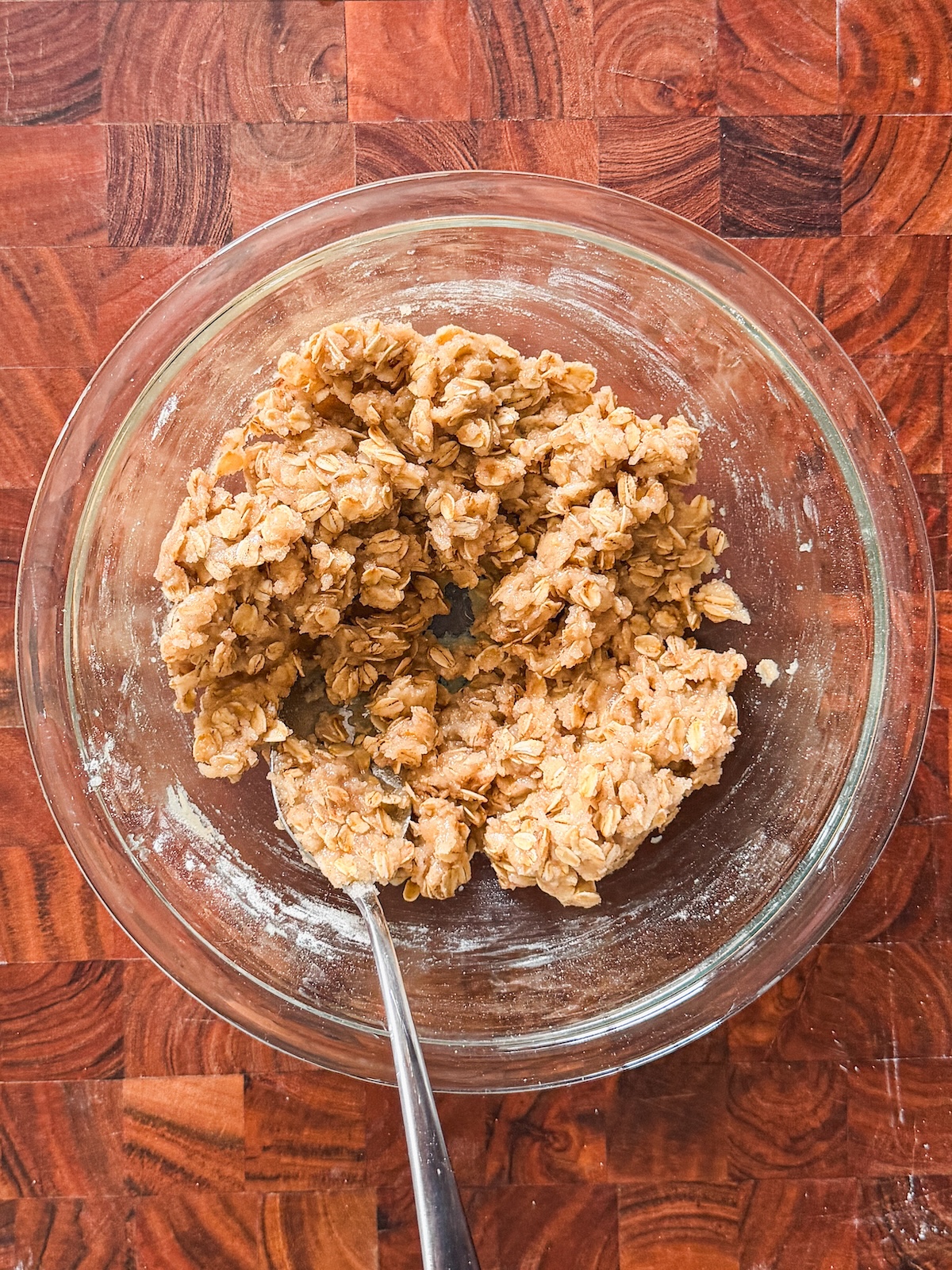 A glass bowl filled with oatmeal crumble dough and a metal spoon, placed on a wooden countertop with a checkered pattern.