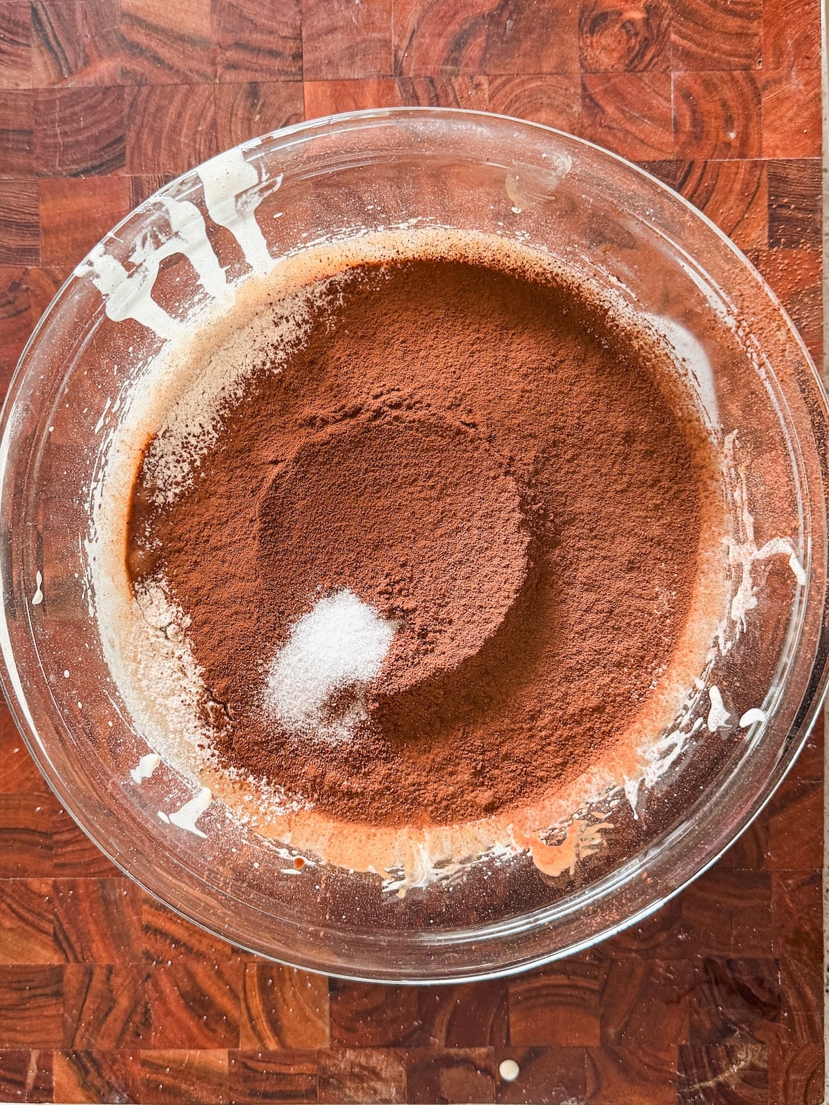 A glass mixing bowl on a wooden surface contains sifted cocoa powder, sugar, and flour, with some ingredients clinging to the sides of the bowl.