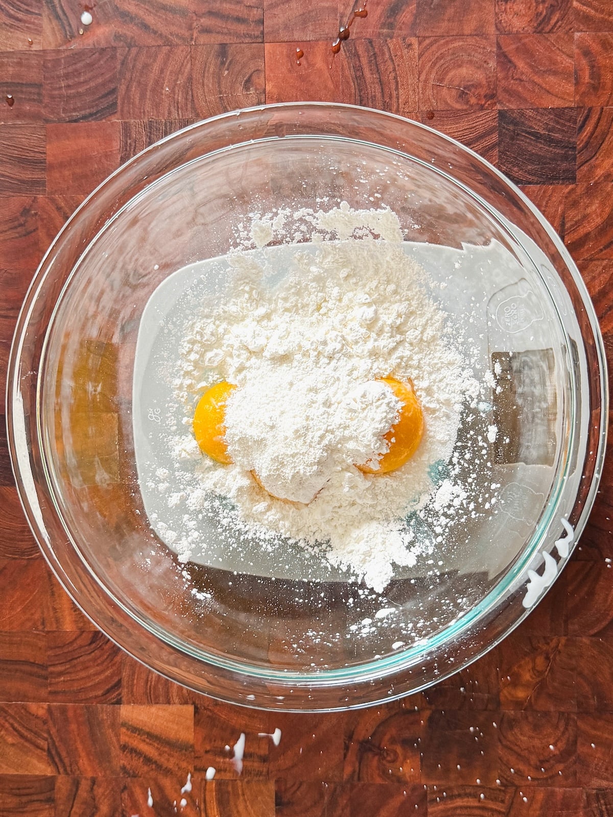 A glass mixing bowl on a wooden surface contains two egg yolks and a heap of cornstarch.