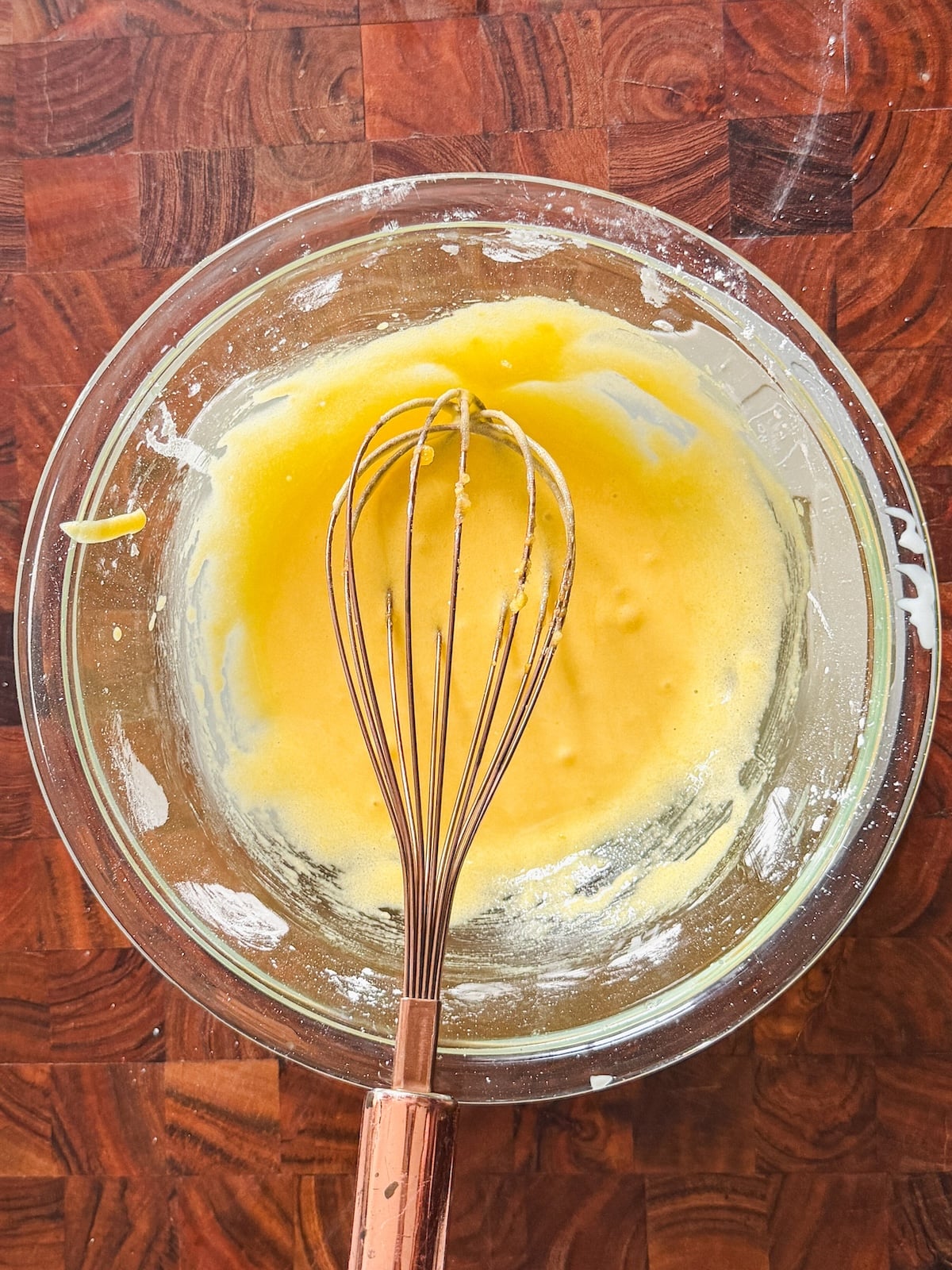 A glass bowl with egg yolks and cornstarch being mixed by a metal whisk, sitting on a wooden countertop. Batter residue is visible on the sides of the bowl.