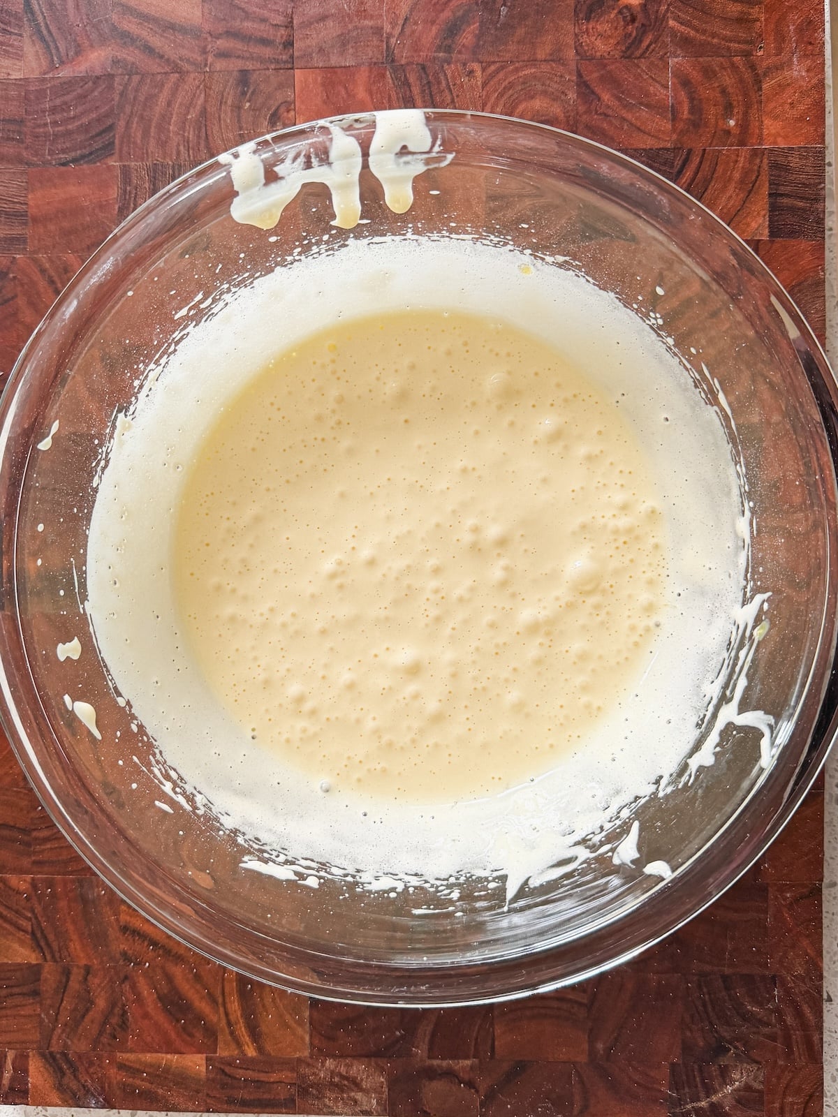 A glass bowl filled with pale, frothy egg and sugar batter sits on a wooden countertop. Some of the batter is splattered on the rim of the bowl.