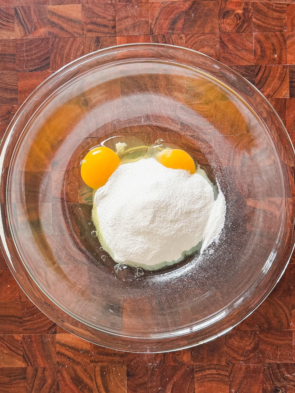 A glass mixing bowl containing two raw eggs and a mound of white sugar, placed on a wooden surface.