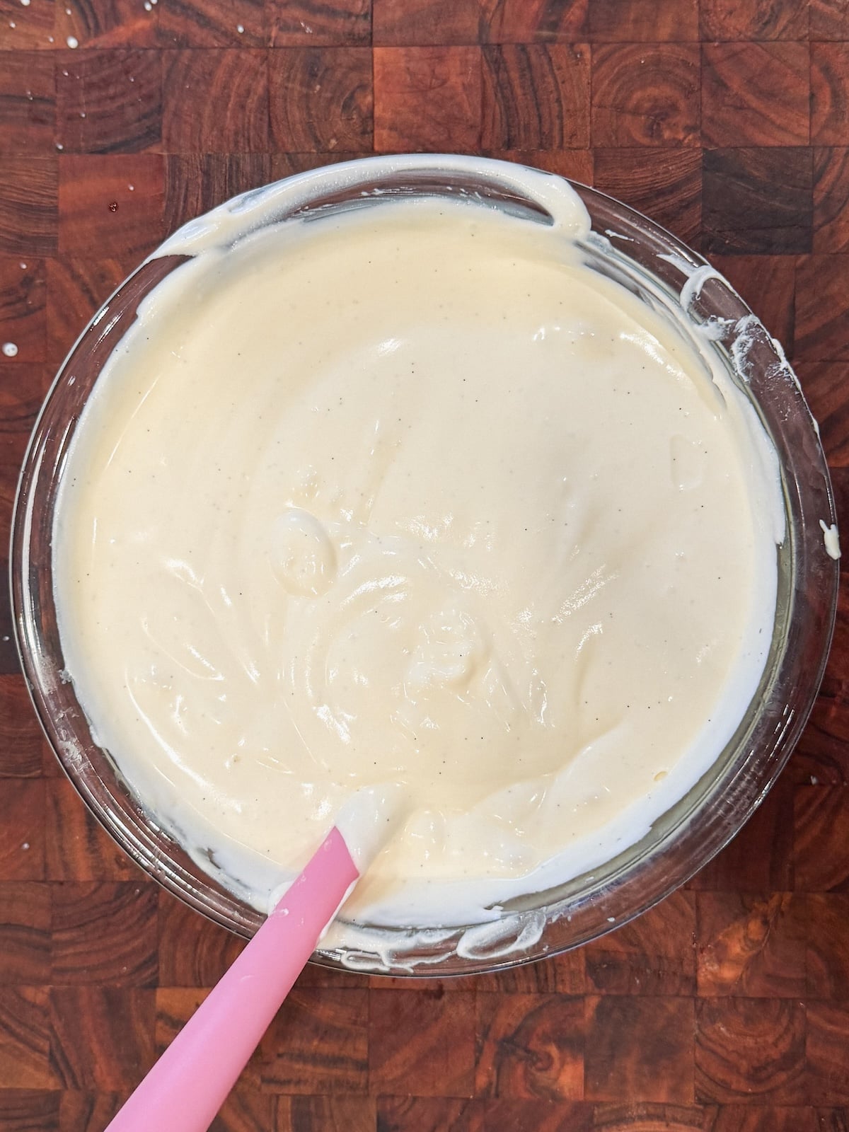 A glass bowl filled with creamy, pale vanilla pudding sits on a wooden countertop. A pink spatula with a white tip rests in the batter, which has been partially stirred.
