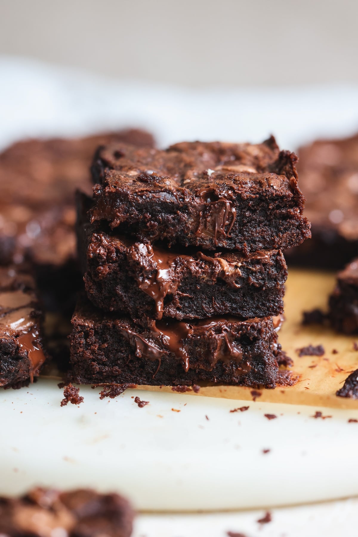 A close-up of three rich, fudgy chocolate brownies stacked on top of each other, with gooey melted chocolate oozing from the middle. More brownies are blurred in the background.