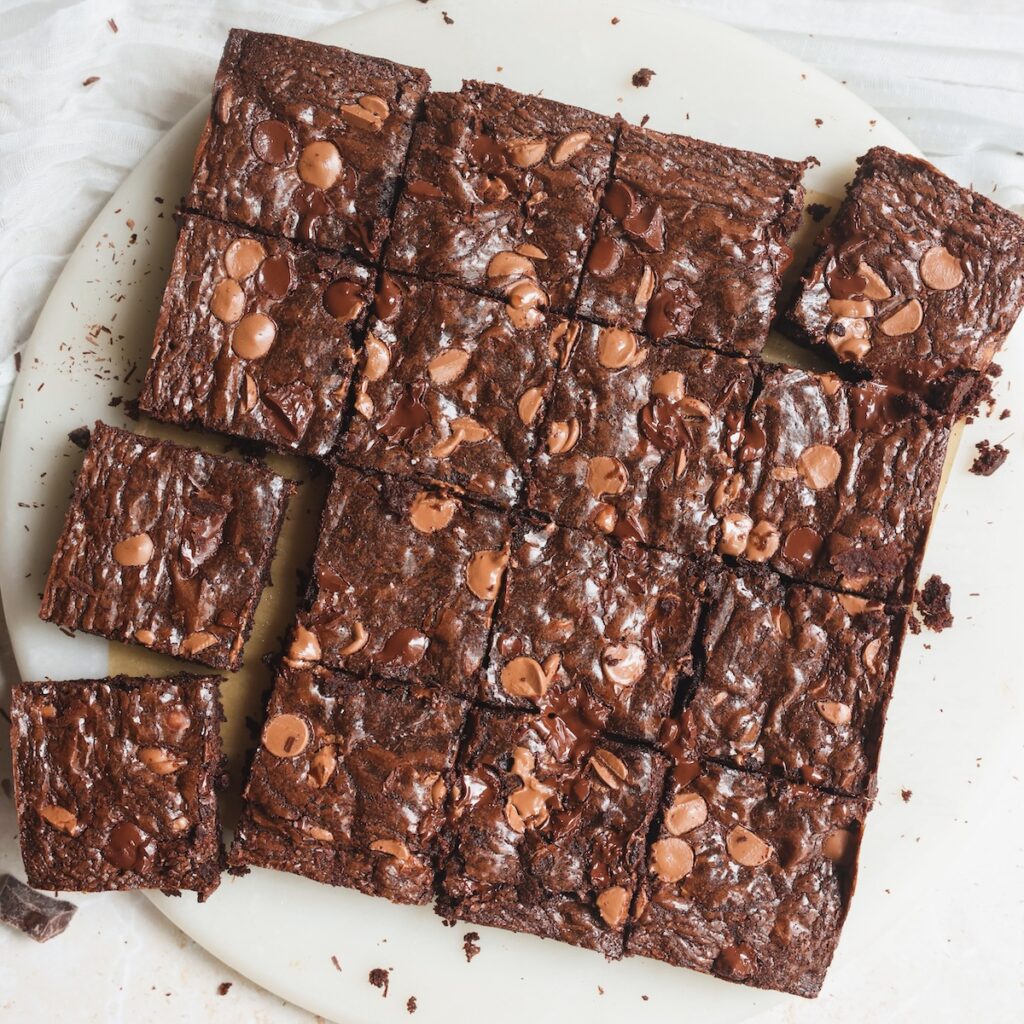 A round white plate topped with sixteen chocolate brownies, each cut into squares and sprinkled with chocolate chips. A few brownies are slightly separated from the group.