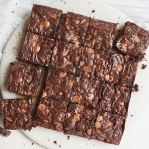 A round white plate topped with sixteen chocolate brownies, each cut into squares and sprinkled with chocolate chips. A few brownies are slightly separated from the group.