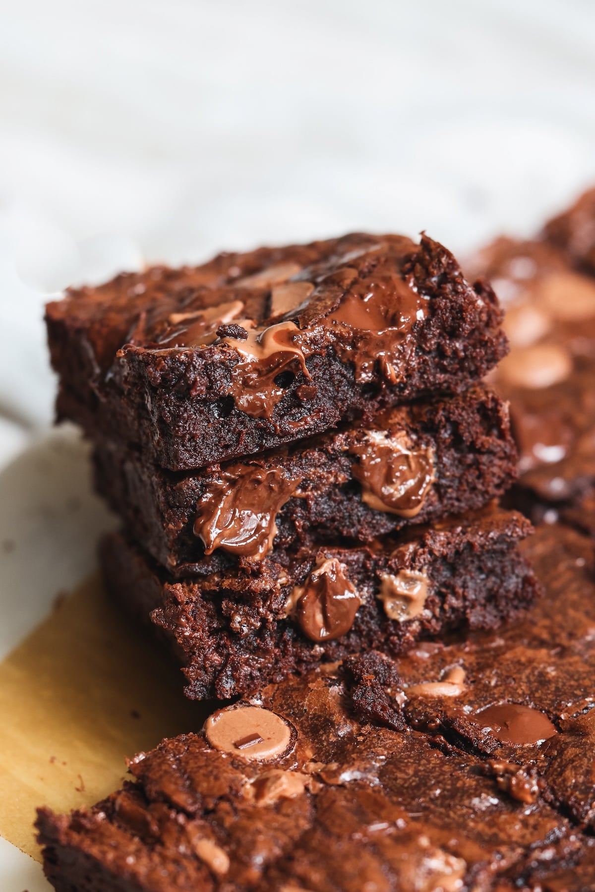 A close-up of a stack of rich, fudgy chocolate brownies with melted chocolate chips, showing gooey texture and a shiny, crackly top. More brownies are visible in the background on parchment paper.