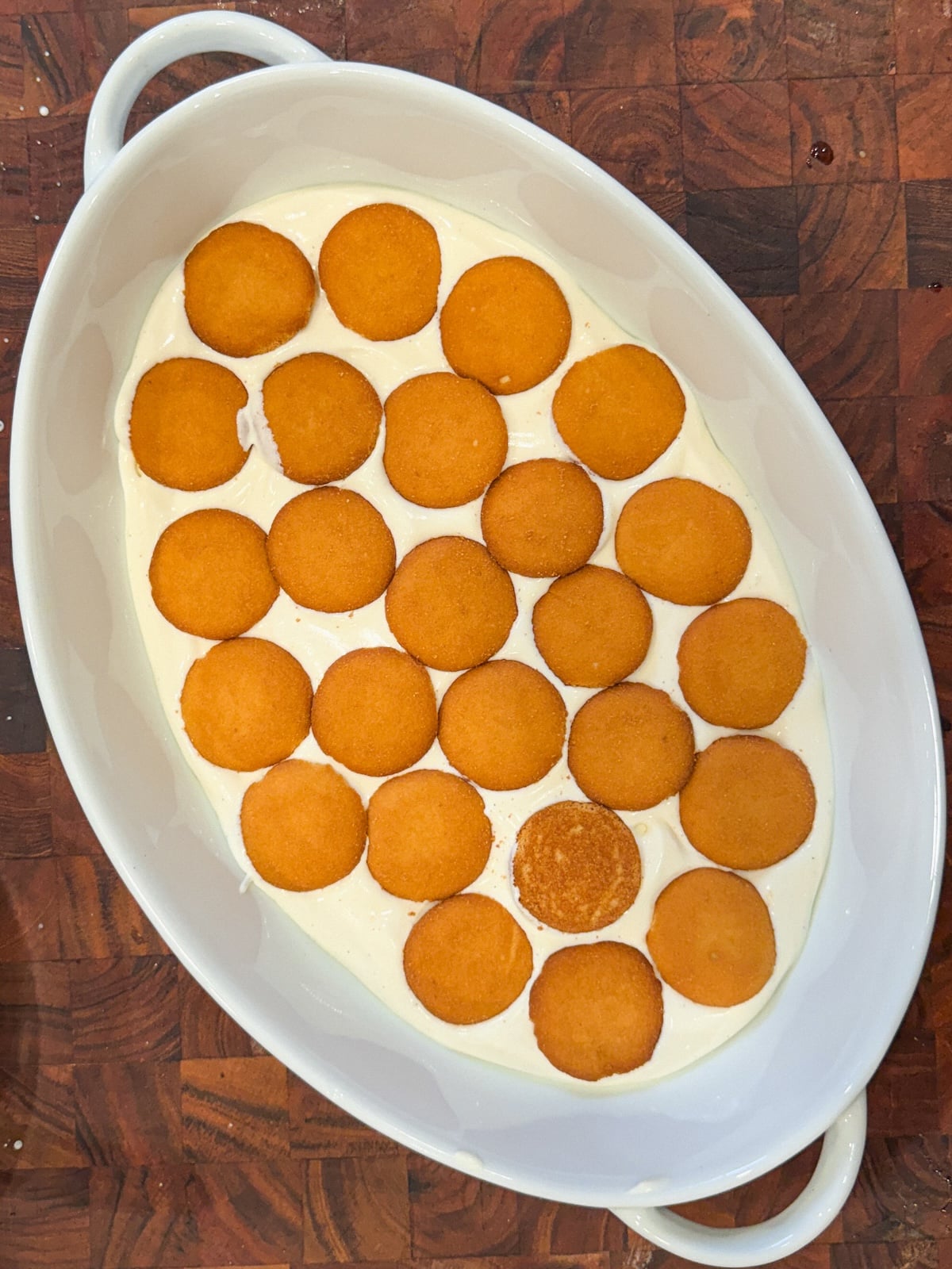 An oval white dish filled with a layer of vanilla pudding topped with evenly arranged round vanilla wafer cookies, placed on a wooden surface.
