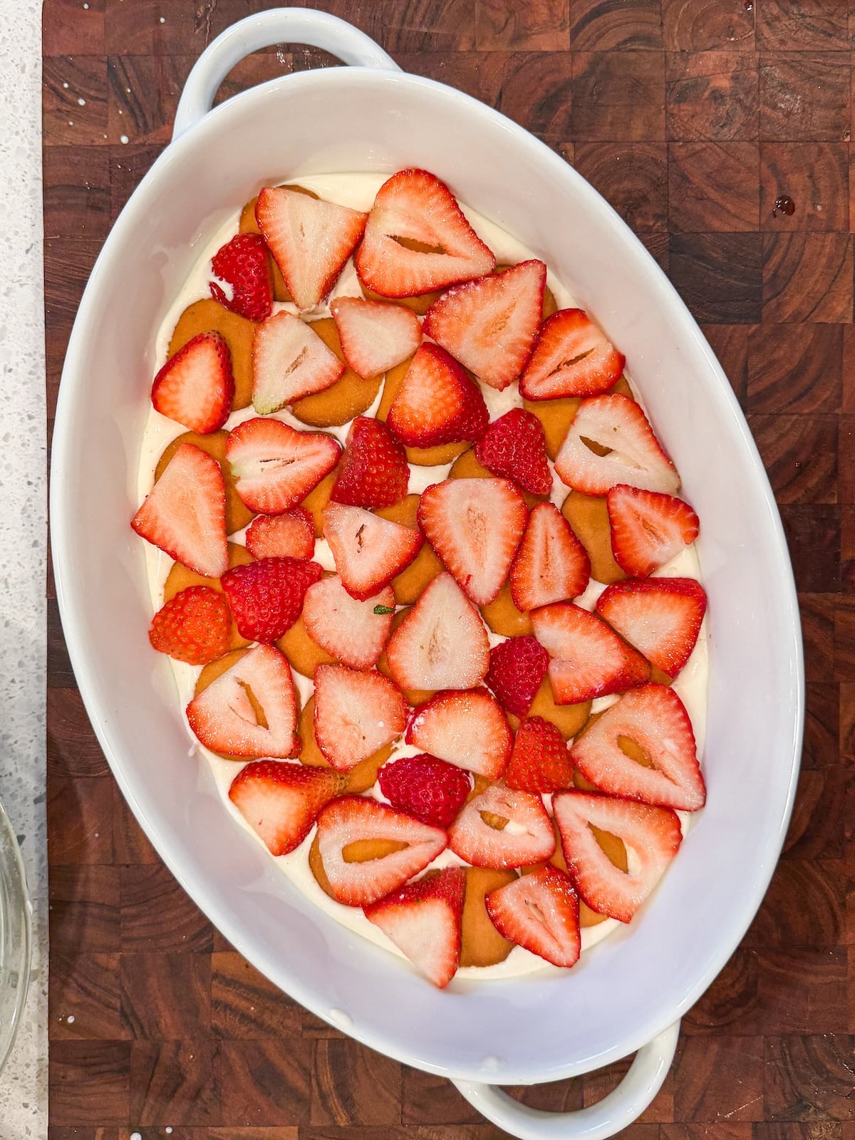 A white oval baking dish filled with layers of sliced strawberries, round vanilla wafer cookies, and a creamy pudding, placed on a wooden surface.