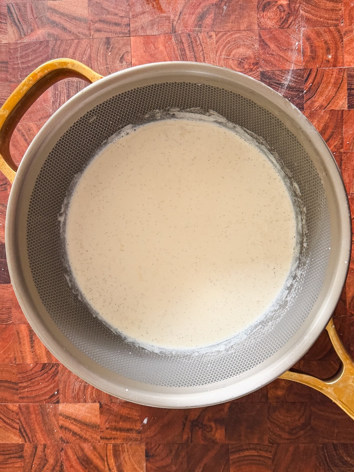 A metal pan with a golden handle contains a mix of milk and cream that is simmering, seen from above, sitting on a reddish-brown wooden surface.