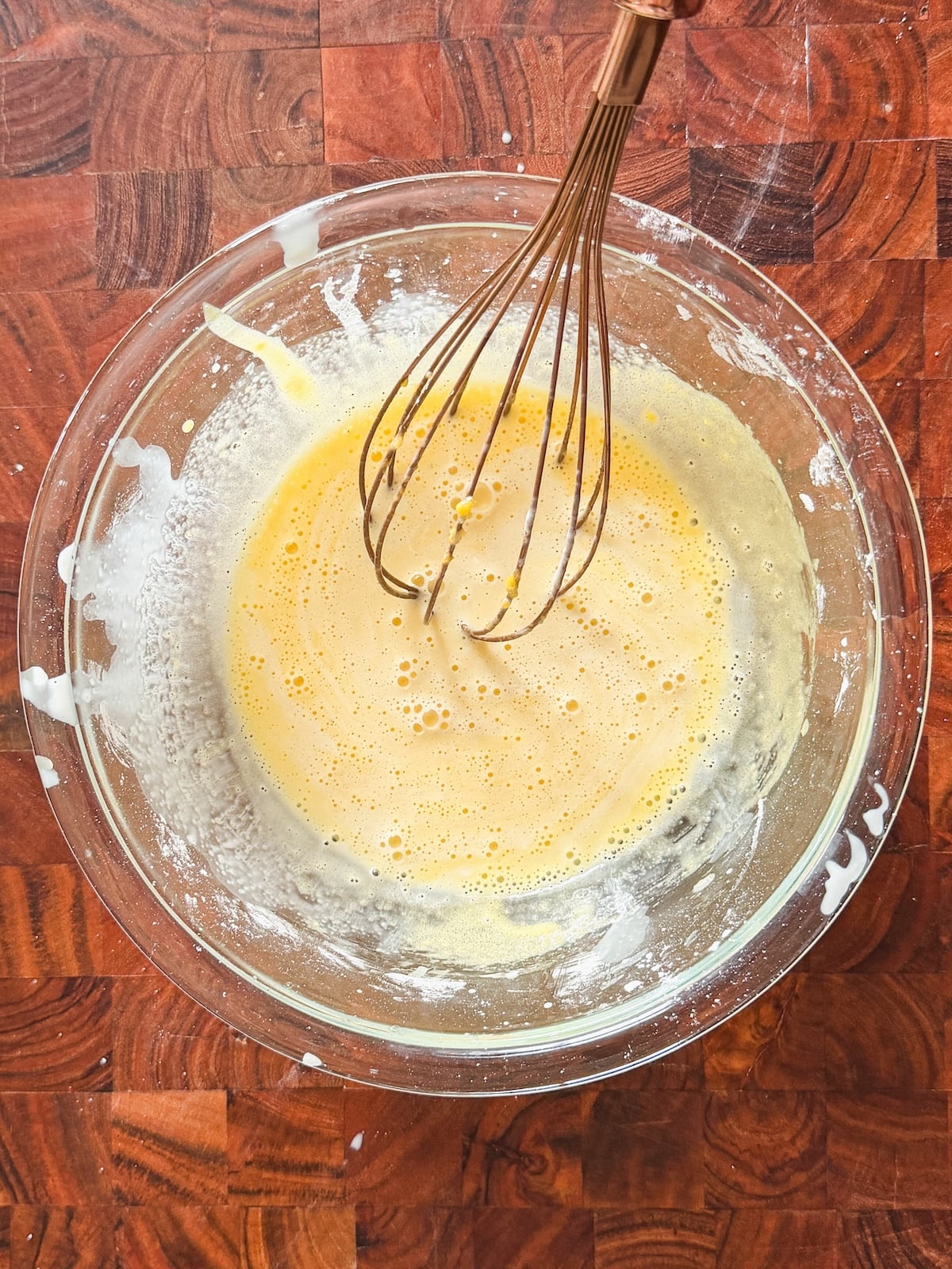 A metal whisk in a large glass bowl containing a pale yellow, frothy mixture sits on a wooden countertop, with some splatters of the mixture visible around the bowl's rim.
