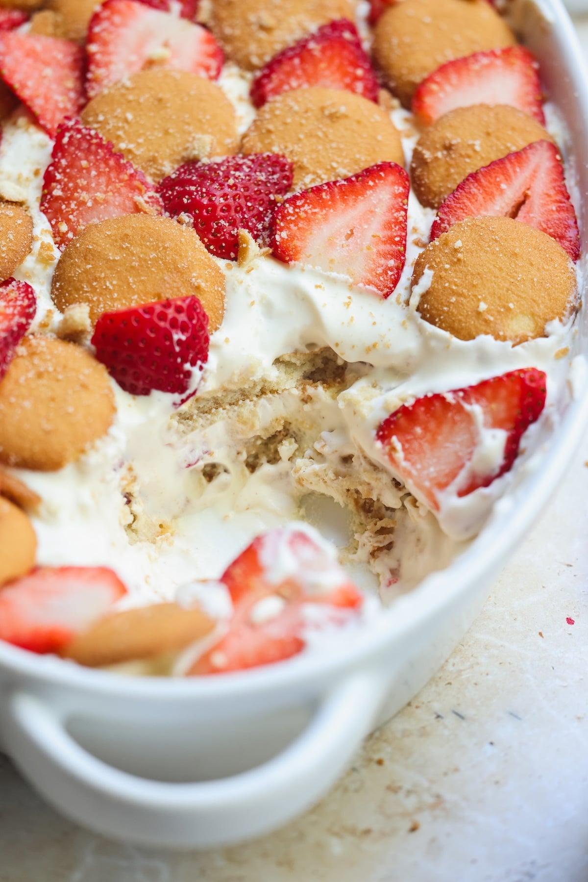 A close-up of a strawberry pudding in a white dish, layered with pudding, vanilla wafers, and fresh strawberry slices. A portion has been scooped out, revealing the creamy layers inside.