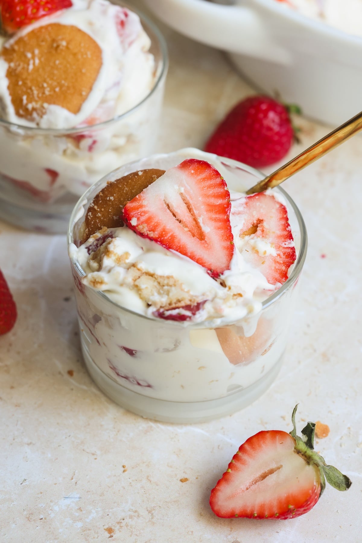 A glass filled with layered dessert of pudding, vanilla wafers, and sliced strawberries, with a spoon and fresh strawberries scattered around on a light surface.