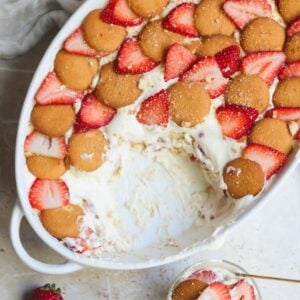 A white oval dish filled with creamy strawberry pudding, topped with vanilla wafer cookies and sliced strawberries. Part of the dessert has been scooped out, revealing luscious layers inside. A strawberry sits nearby.