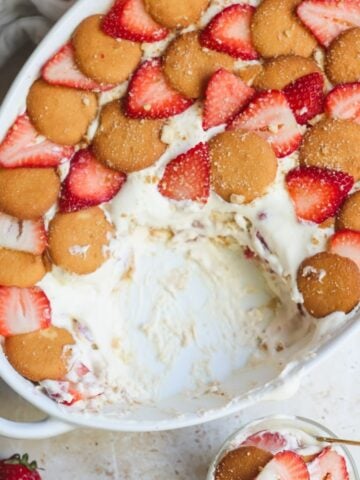 A white oval dish filled with creamy strawberry pudding, topped with vanilla wafer cookies and sliced strawberries. Part of the dessert has been scooped out, revealing luscious layers inside. A strawberry sits nearby.