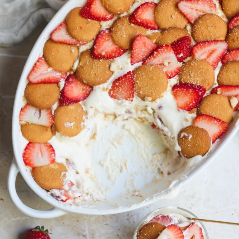 A white oval dish filled with creamy strawberry pudding, topped with vanilla wafer cookies and sliced strawberries. Part of the dessert has been scooped out, revealing luscious layers inside. A strawberry sits nearby.