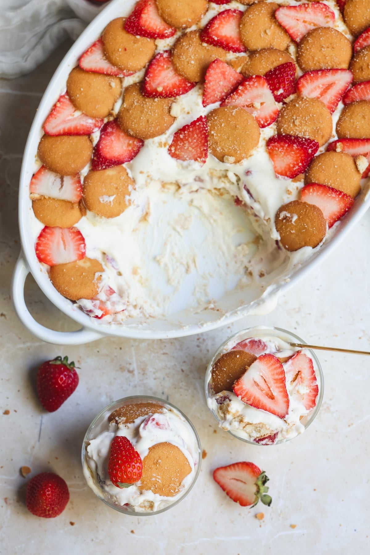 A baking dish of layered strawberry pudding with vanilla wafers, sliced strawberries, and creamy filling, some servings scooped out. Two small glasses with the same dessert sit beside the dish, garnished with strawberries and wafers.