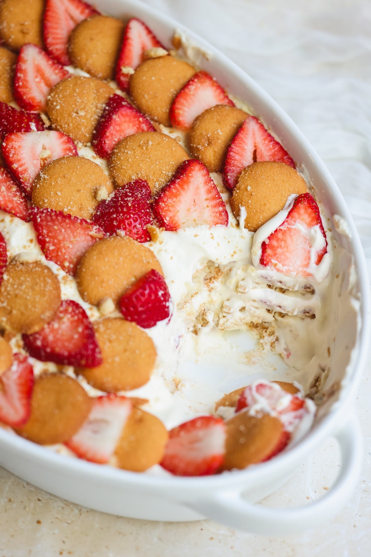 A close-up of a dessert dish with layers of vanilla wafers, sliced strawberries, and creamy filling, with a portion scooped out showing the layered texture inside a white oval casserole dish.