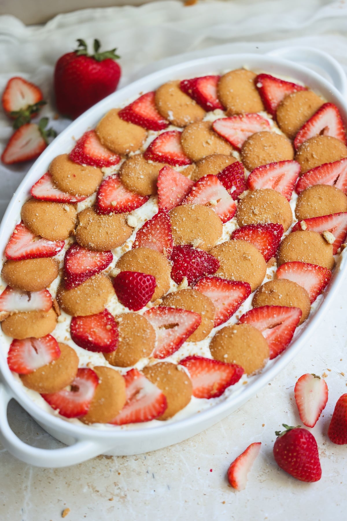 A white oval dish filled with a layered dessert topped with sliced strawberries and round vanilla wafer cookies, with whole strawberries and crumbs scattered nearby on a white surface.