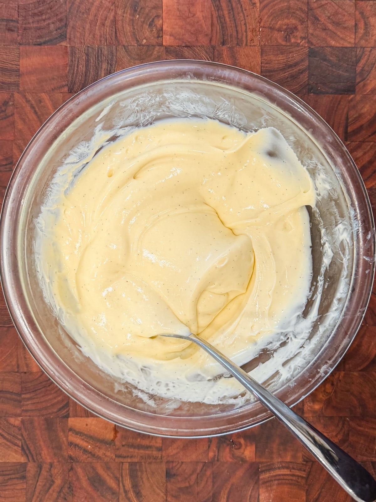 A glass bowl filled with creamy, thick pale yellow pudding sits on a wooden surface. A metal spoon is resting in the mixture, which appears smooth and thick.