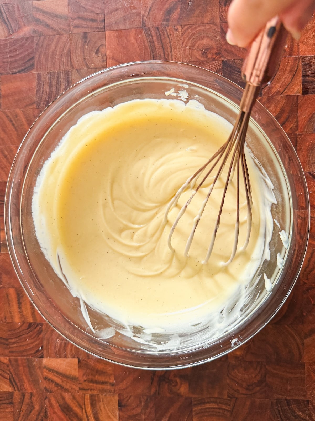 A hand using a whisk to mix creamy batter in a clear glass bowl, placed on a wooden surface.