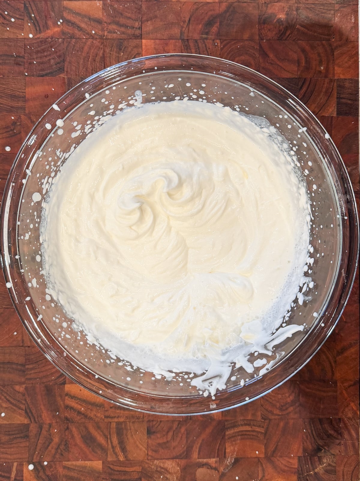 A glass bowl filled with freshly whipped cream sits on a wooden cutting board. The cream has soft peaks and a light, airy texture, with some splatters around the edges of the bowl.