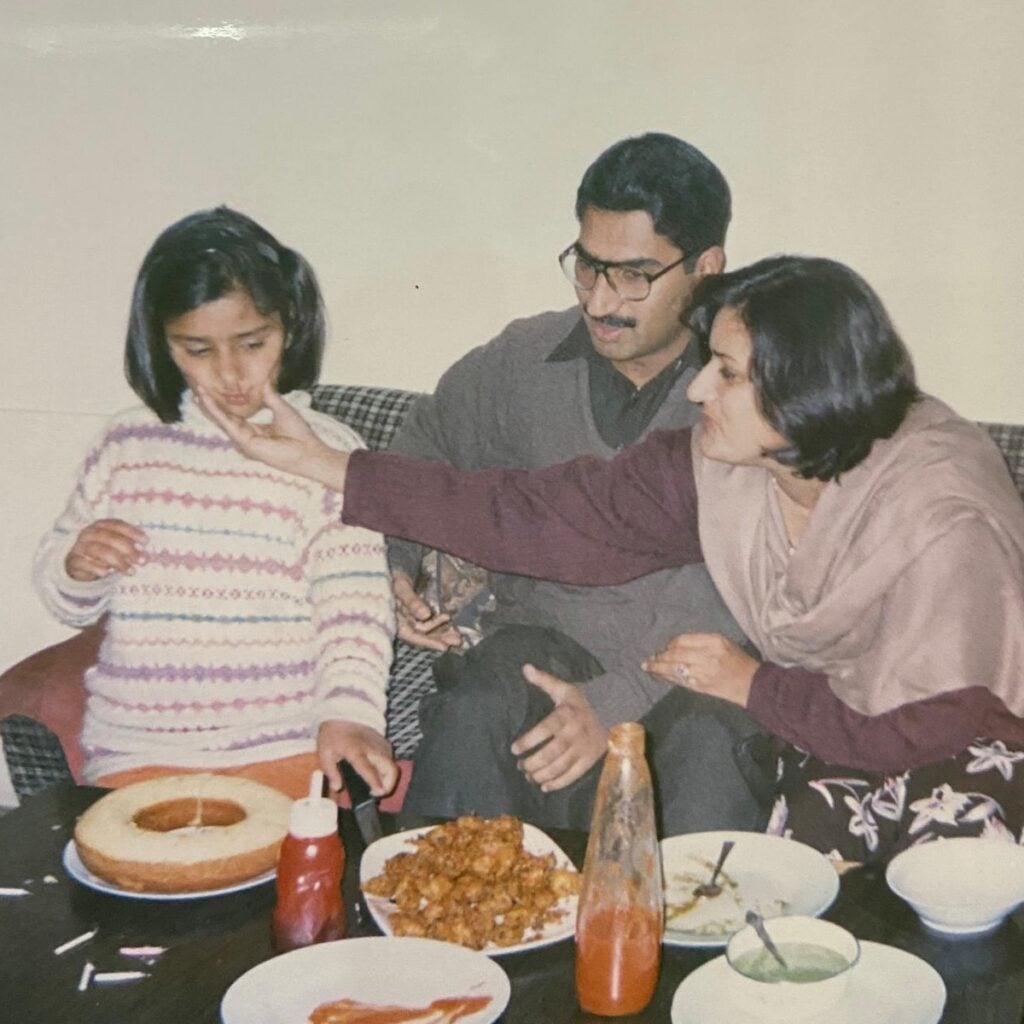 A woman gently feeds a young girl beside a man on a sofa. The table in front of them has a cake, fried snacks, ketchup, and dishes. They appear to be sharing a meal in a cozy home setting.