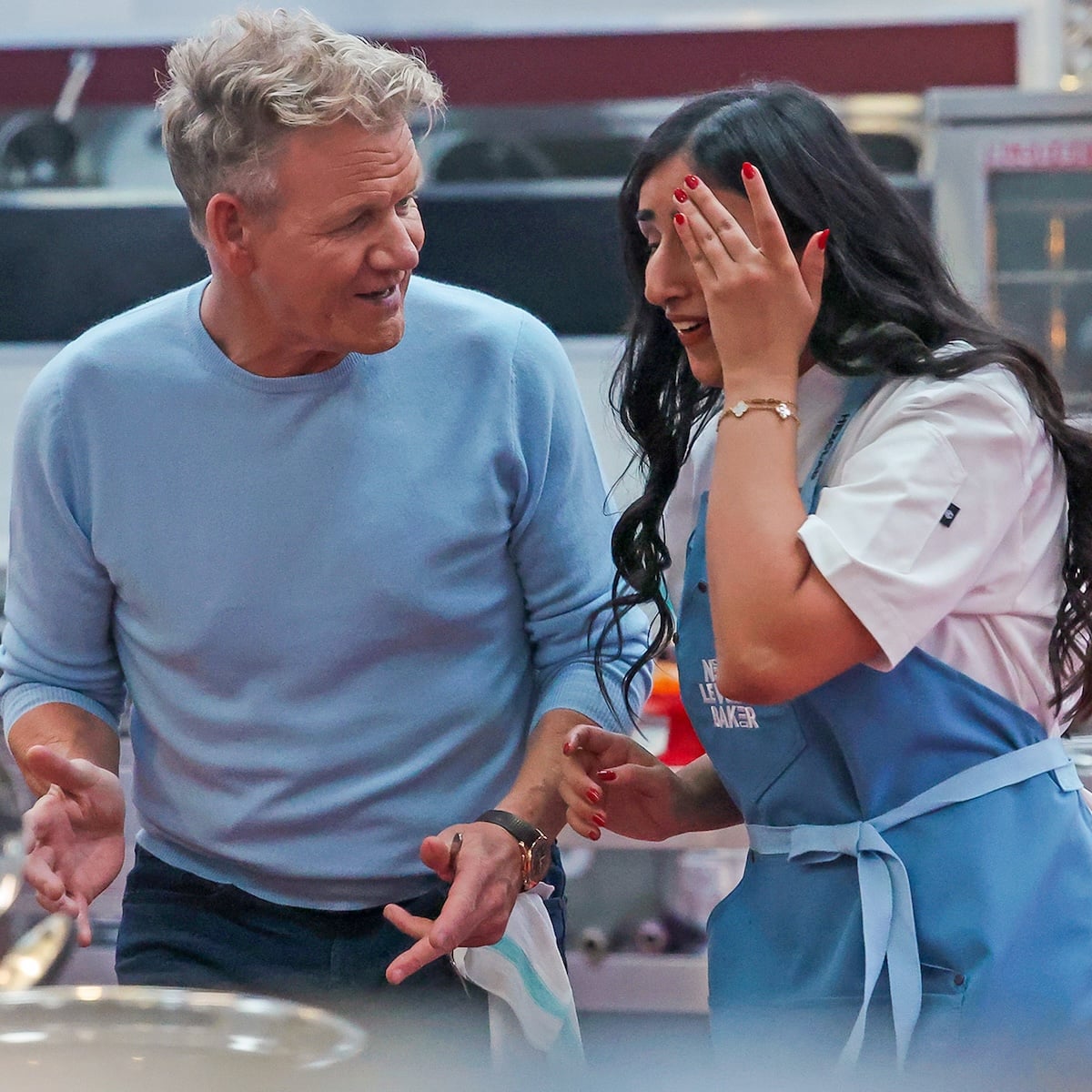 A man in a light blue sweater talks animatedly to a woman in a blue apron, who smiles and covers one eye with her hand, in what appears to be a busy kitchen setting.