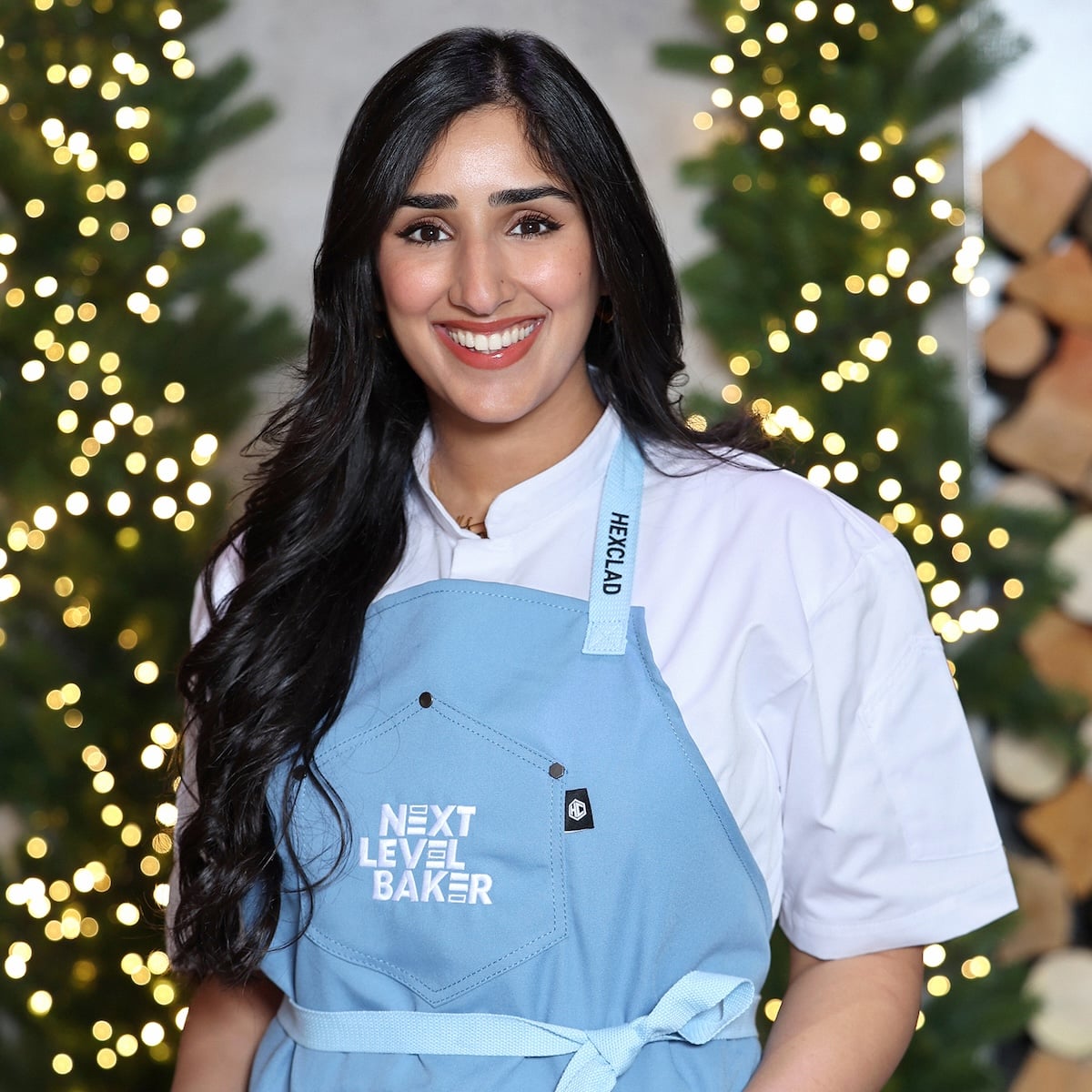A smiling woman with long dark hair wearing a blue apron that says NEXT LEVEL BAKER stands in front of decorated trees with string lights and stacked firewood in the background.