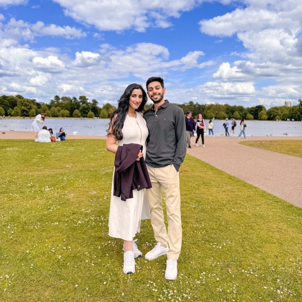 A smiling couple stands together on a grassy area by a lake in a park, with people in the background under a blue sky with scattered clouds. The woman wears a white dress; the man wears beige pants and a dark top.