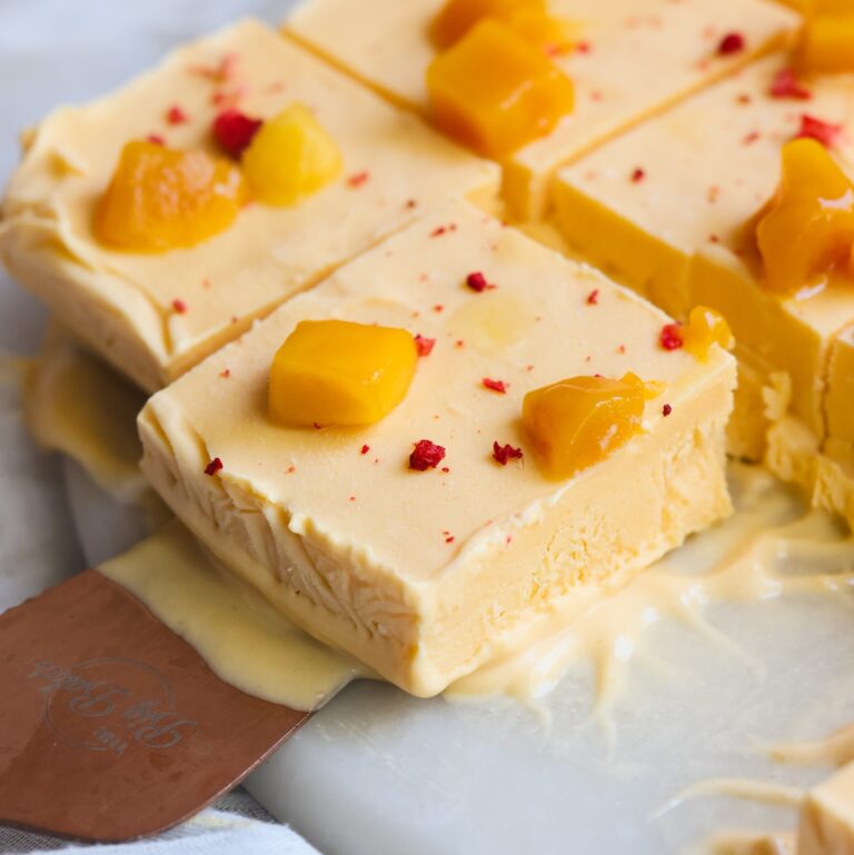 Several creamy, yellow mango kulfi ice cream bars topped with diced mango and red bits are sliced and served on a white surface. One bar is being lifted with a metal spatula. Some melting is visible around the bars.
