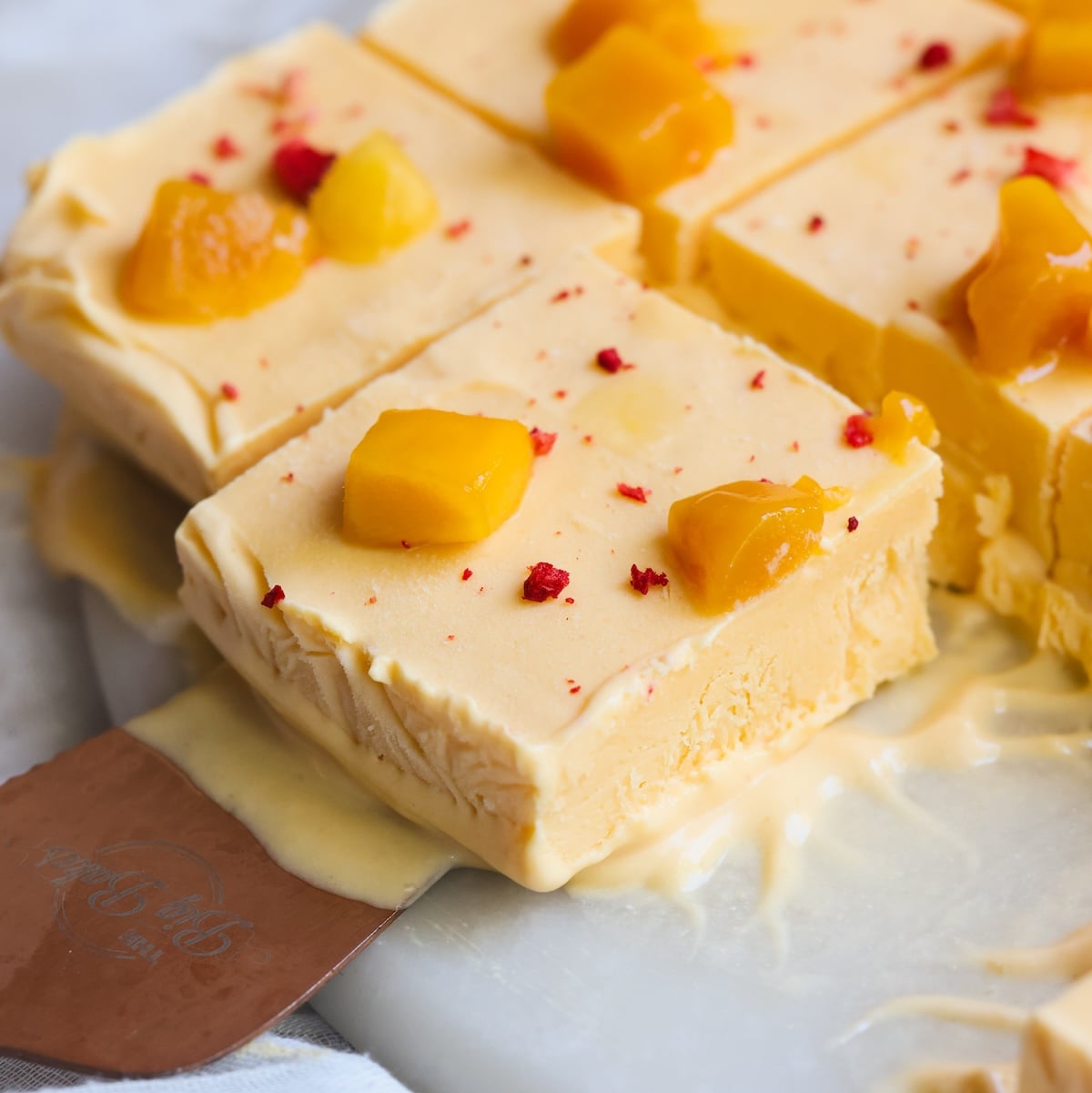 Several creamy, yellow mango kulfi ice cream bars topped with diced mango and red bits are sliced and served on a white surface. One bar is being lifted with a metal spatula. Some melting is visible around the bars.