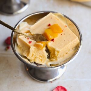 A metal bowl filled with creamy mango kulfi ice cream, topped with small mango pieces. A spoon is scooping out a piece of the ice cream. The bowl sits on a light-colored surface.