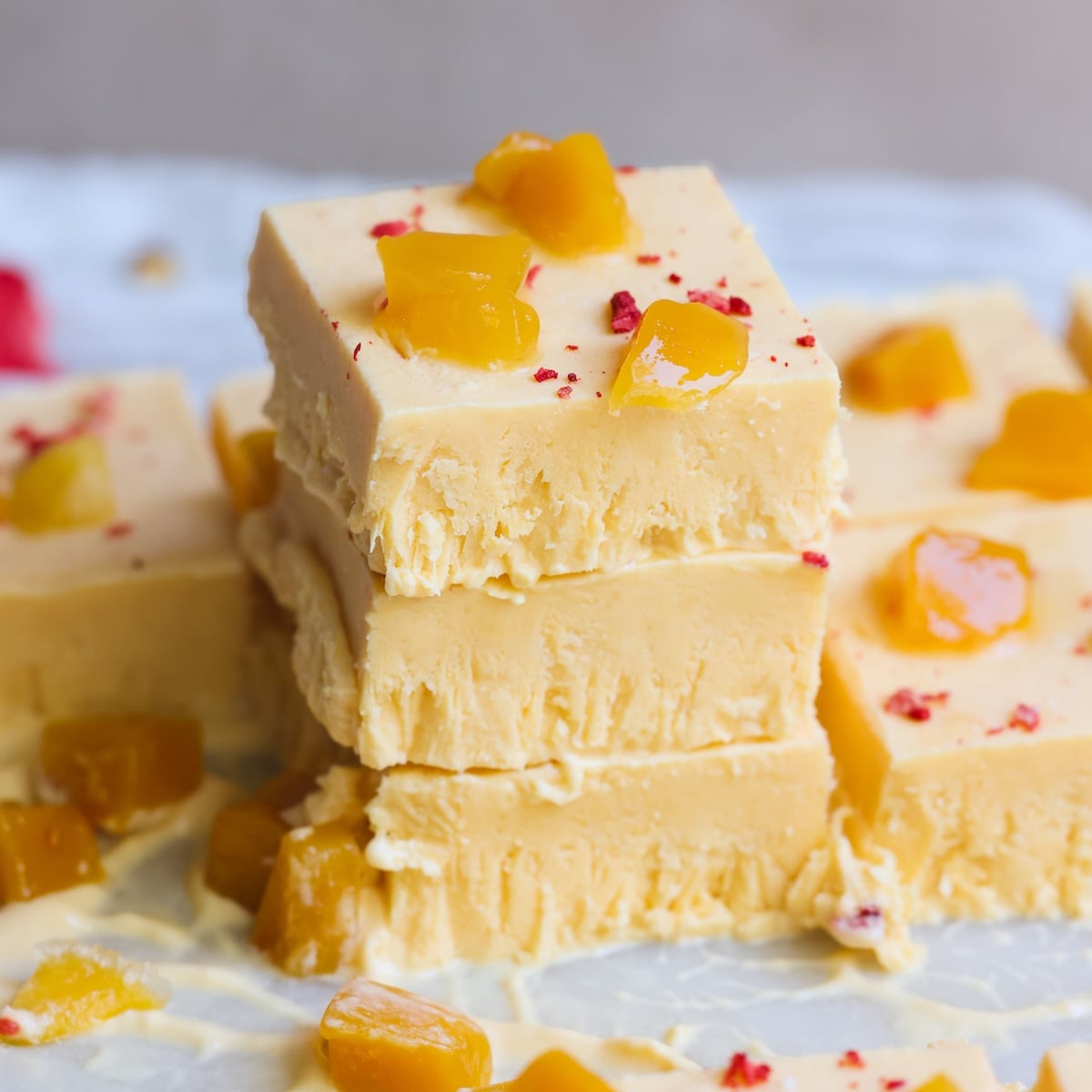 Three creamy yellow mango kulfi slices are stacked, topped with small diced mango pieces and red sprinkles. More slices and mango pieces are scattered around on a white surface.