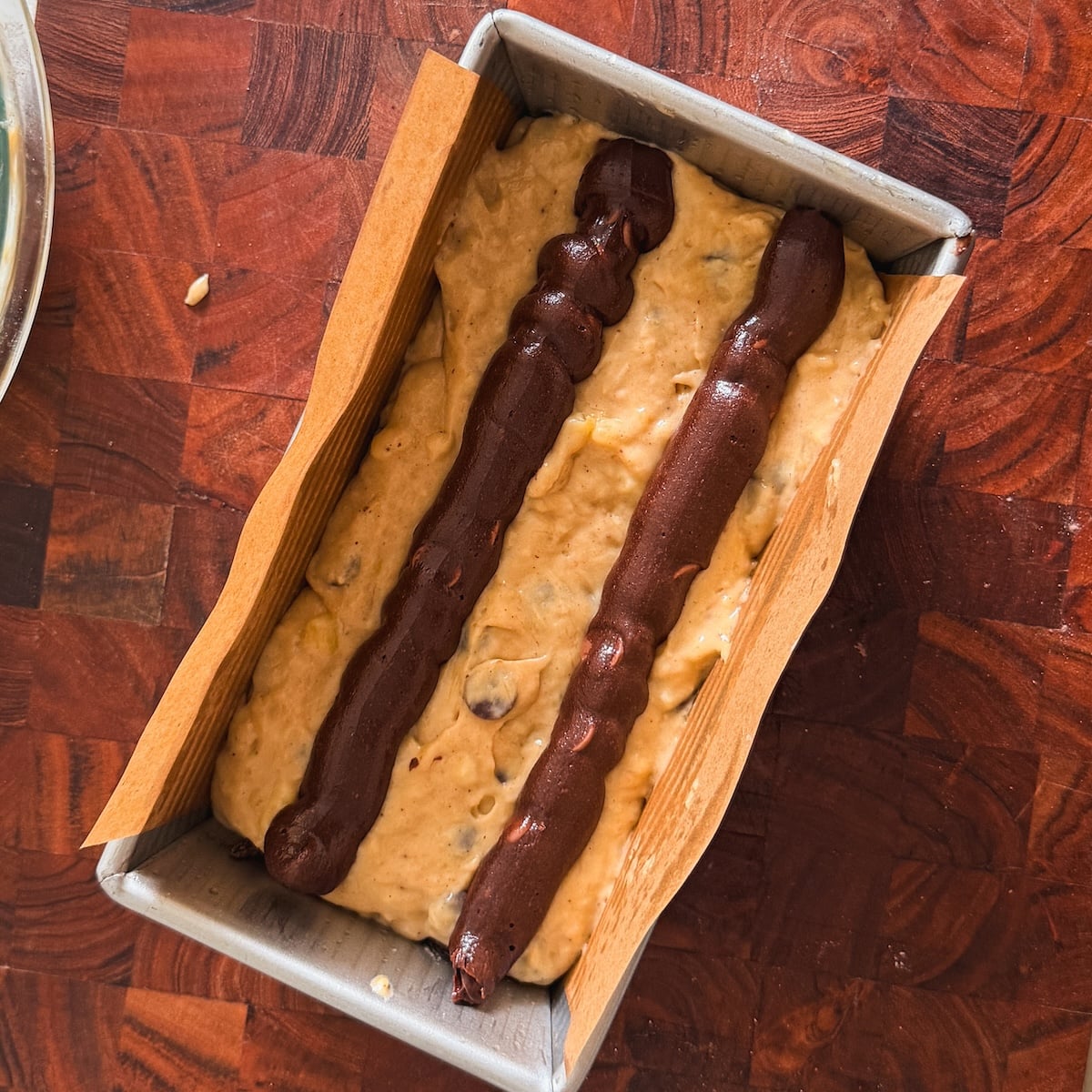 A loaf pan lined with parchment paper contains unbaked banana bread batter with two lines of brownie batter on top, resting on a wooden surface.