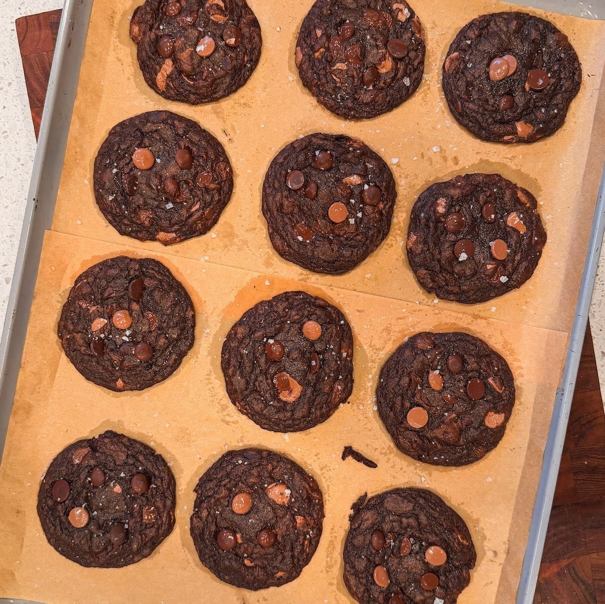 A baking tray lined with parchment paper holds twelve large, round double chocolate chip cookies, each topped with chocolate chips and chunks.