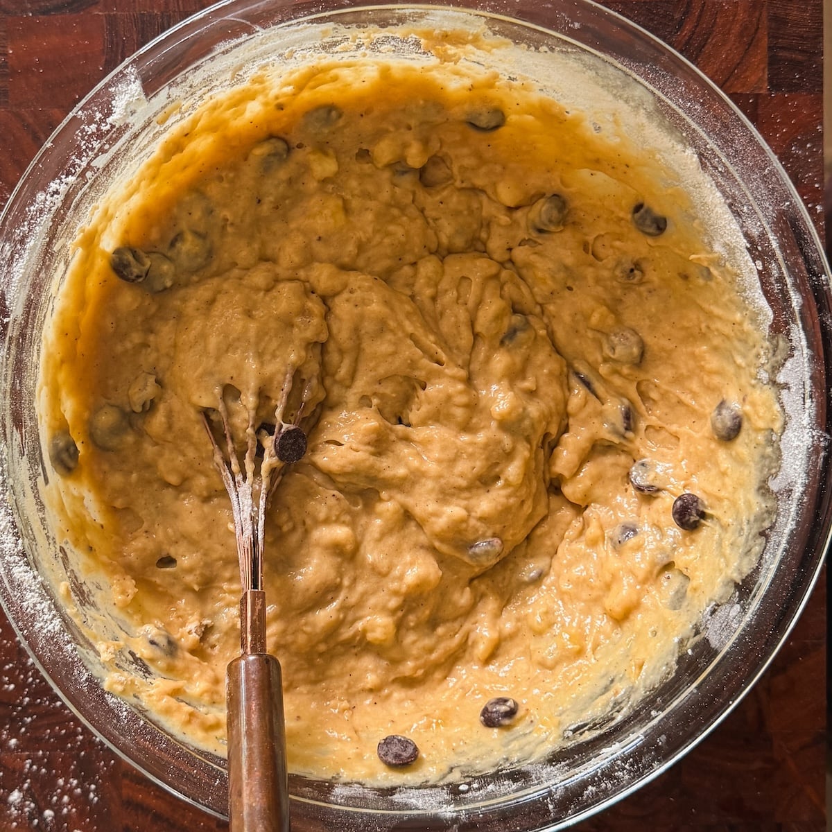 A glass bowl filled with thick banana bread batter mixed with chocolate chips. A whisk with a wooden handle is resting in the batter, and some flour is visible on the bowl's edges.