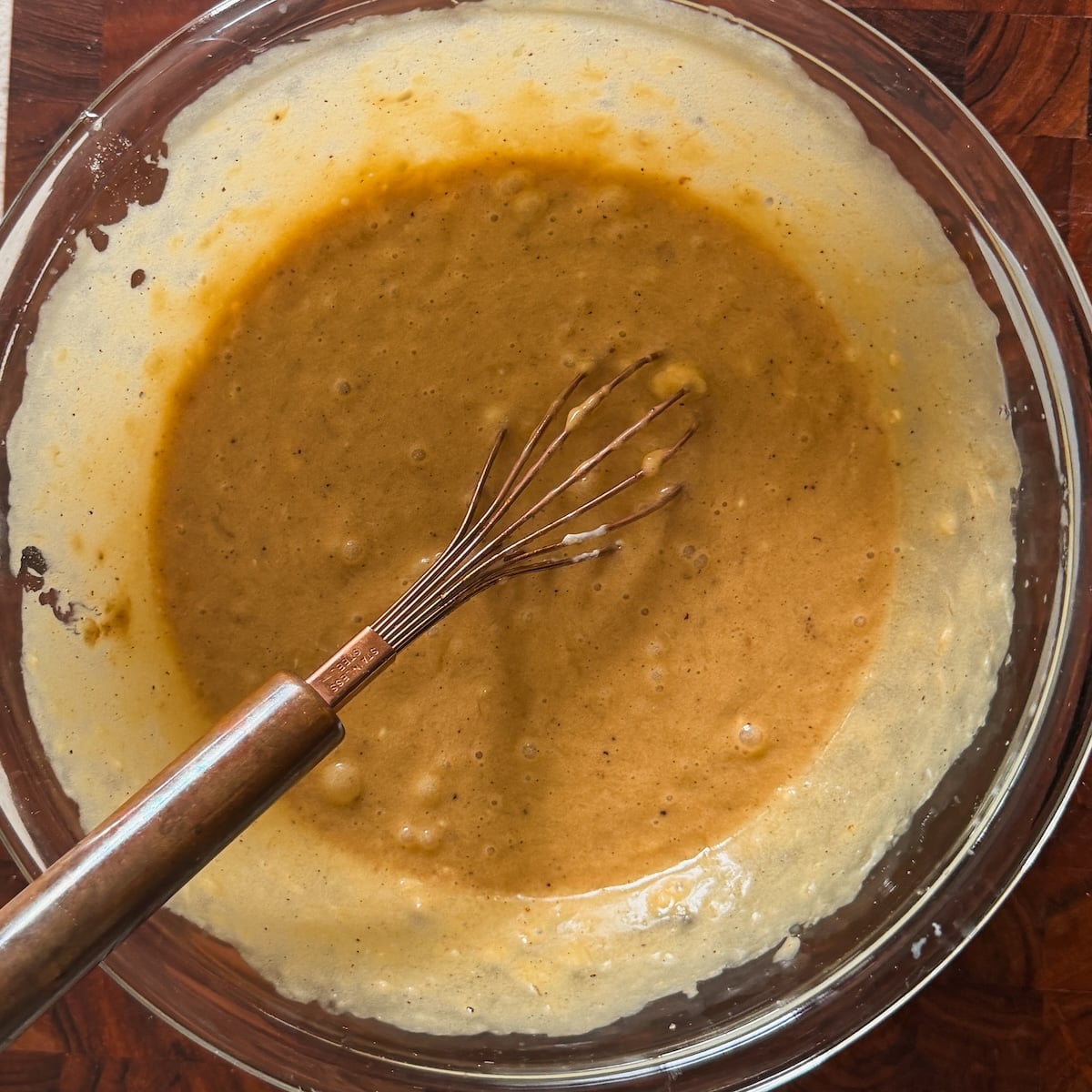 A metal whisk rests in a glass bowl filled with light brown batter, sitting on a dark wooden surface. The batter appears thick and slightly textured, suggesting its being mixed for baking.