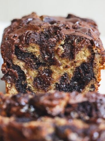 A close-up of a sliced loaf of chocolate brownie swirl banana bread, showing moist, marbled layers of banana bread and melted chocolate. The loaf is set on a light background, and the texture appears rich and gooey.