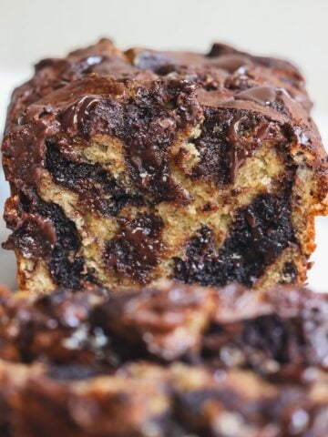 A close-up of a sliced loaf of chocolate brownie swirl banana bread, showing moist, marbled layers of banana bread and melted chocolate. The loaf is set on a light background, and the texture appears rich and gooey.