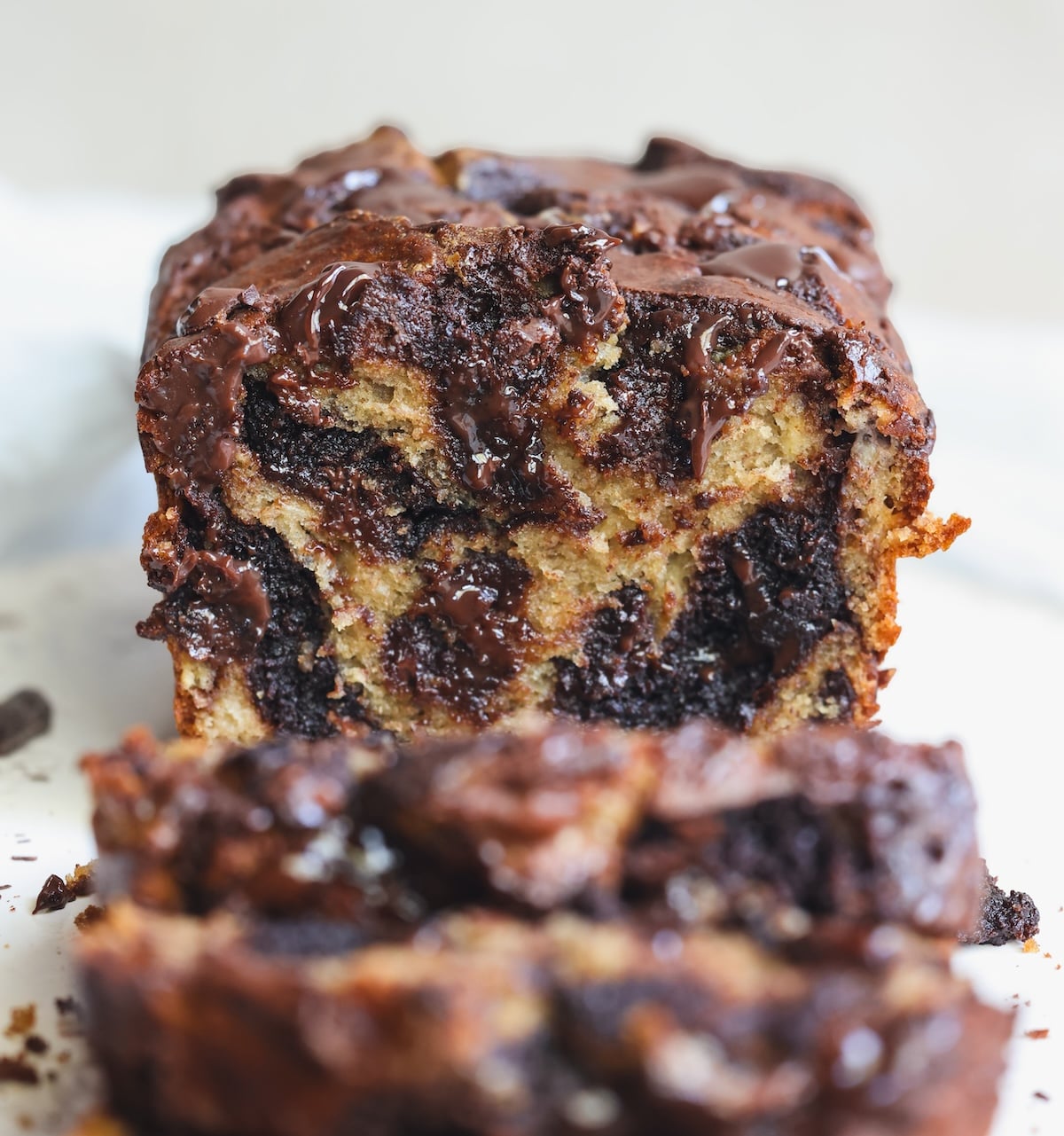 A close-up of a sliced loaf of chocolate brownie swirl banana bread, showing moist, marbled layers of banana bread and melted chocolate. The loaf is set on a light background, and the texture appears rich and gooey.