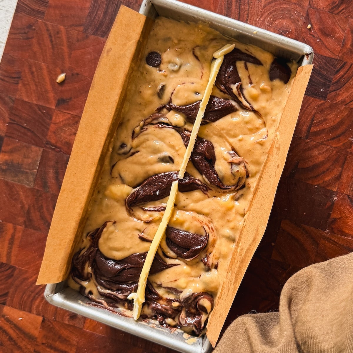 A loaf pan lined with parchment paper is filled with swirled banana bread batter and brownie batter, ready to bake. The pan sits on a wooden surface, and a hand holding an oven mitt is visible in the corner.