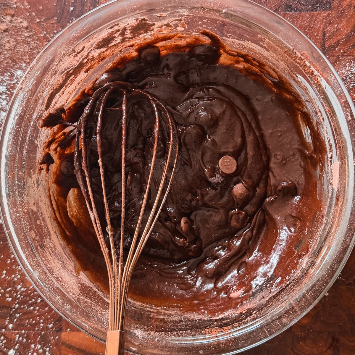 A glass bowl filled with thick chocolate brownie batter and chocolate chips, with a whisk resting inside. The bowl sits on a wooden surface sprinkled with flour.