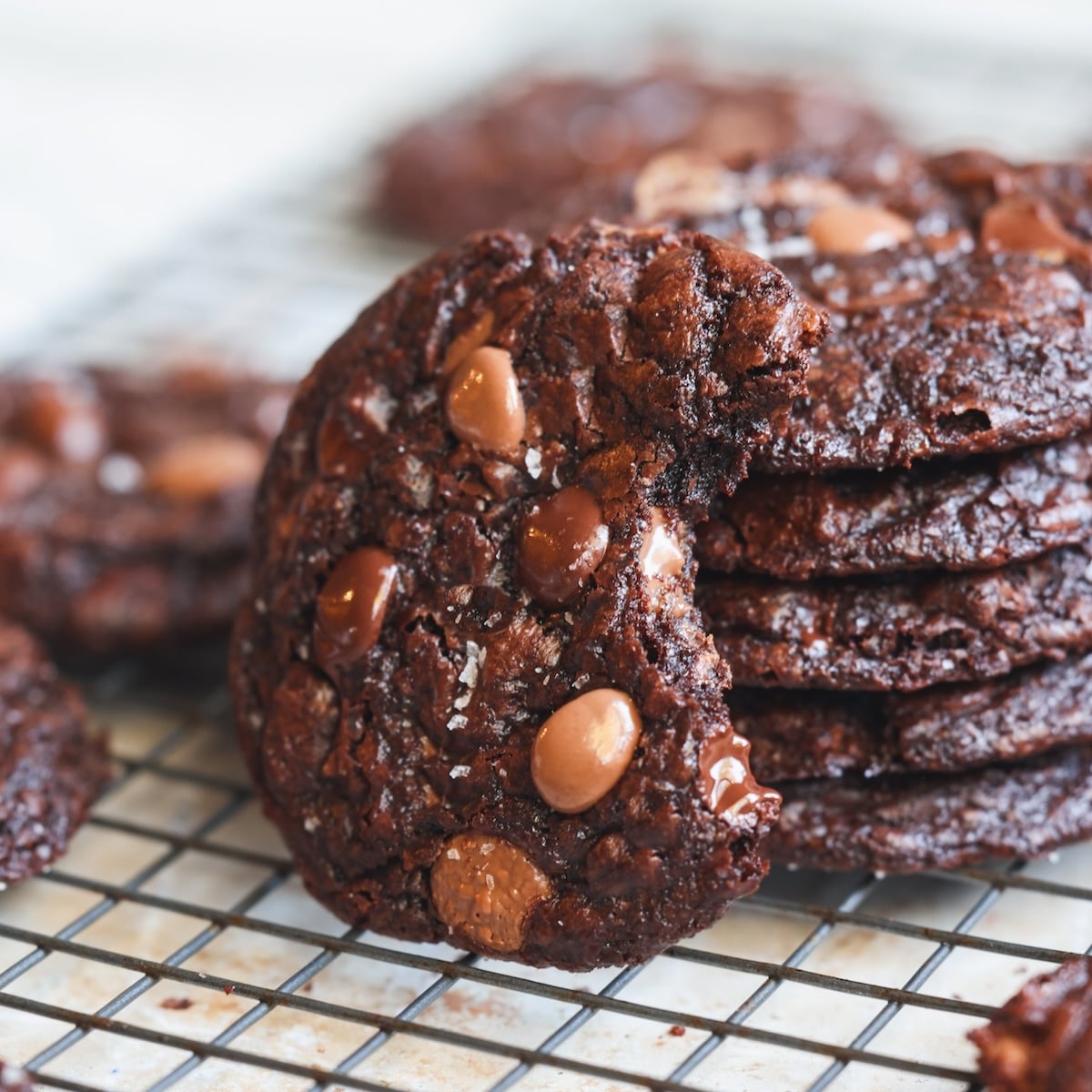A stack of rich, chocolate chocolate-chip cookies on a cooling rack, with the front cookie showing a bite taken out of it. The cookies appear soft, gooey, and studded with melty chocolate chips.