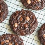 A close-up of chocolate cookies with chocolate chips cooling on a wire rack. The cookies are round, dark, and have a soft texture with several chocolate chips on top.